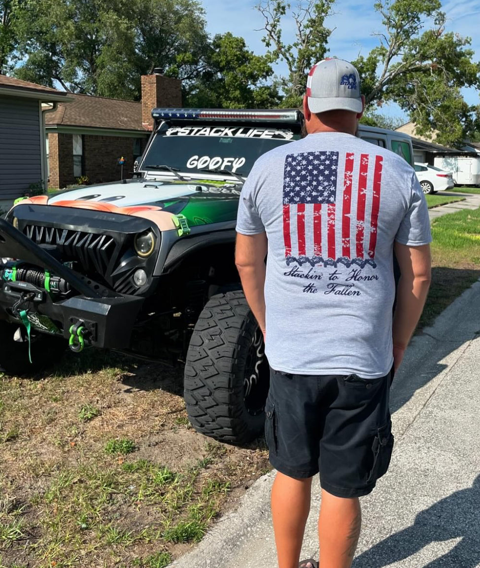 Light grey t shirts with american flag on the back running vertical down back. At the bottom of the flag is a stack of 7 off road vehicles with the words Stackin' to honor the fallen underneath