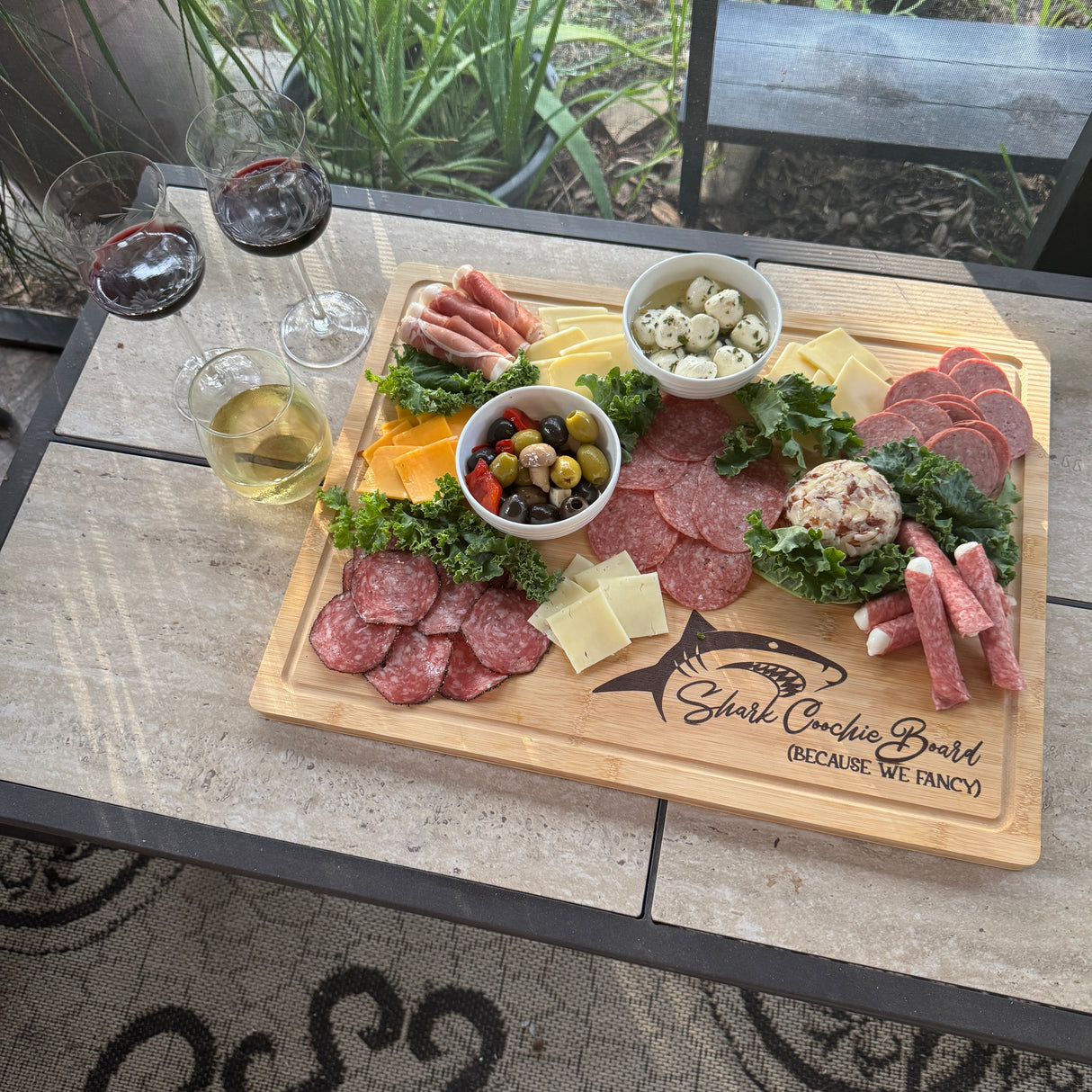 Wooden charcuterie board with meats, cheeses, and olives on a patio table with glasses of wine.