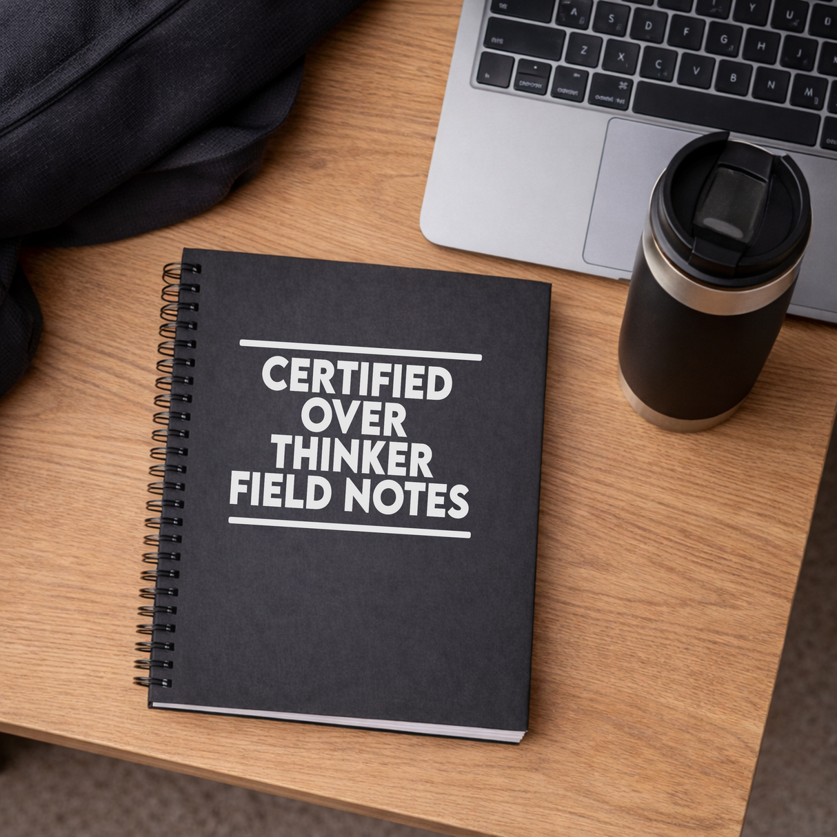 black spiral bound notebook with certified overthinker field notes on the front of it. image is on a desk with a laptop and coffee mug