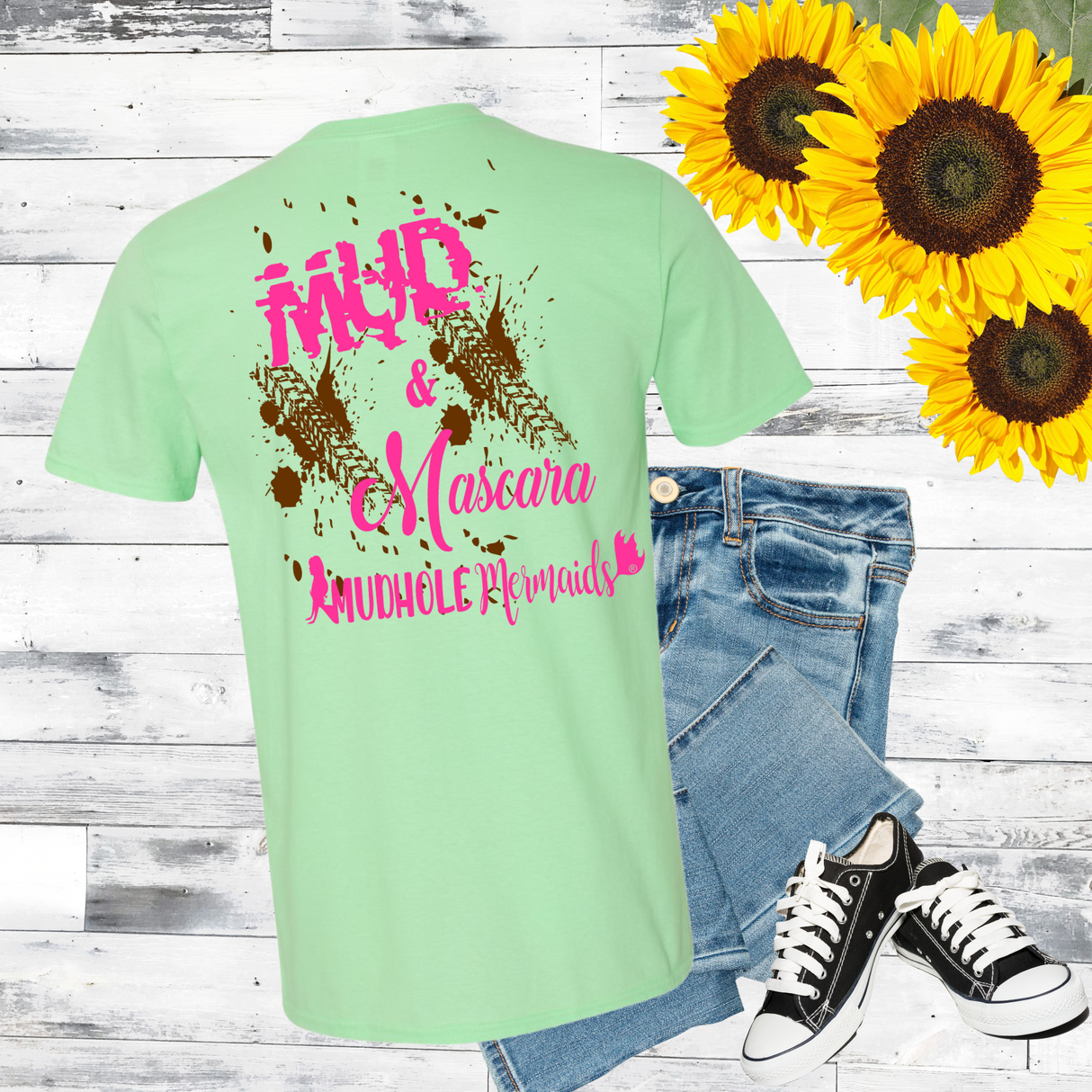 Light green t-shirt with pink text, jeans, and sneakers on a wooden surface with sunflowers. Mud And Mascara logo in pink