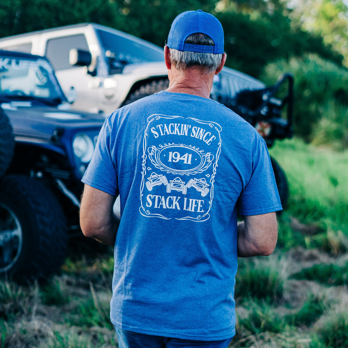 A person wearing a blue t-shirt with 'STACKIN' SINCE 1941' printed on the front, standing next to vehicles in a grassy area.