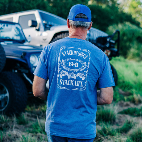 A person wearing a blue t-shirt with 'STACKIN' SINCE 1941' printed on the front, standing next to vehicles in a grassy area.